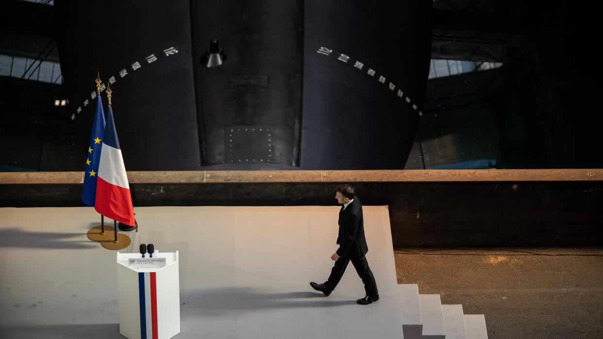 Emmanuel Macron, President of France, walks toward a lectern at the Île Longue military base in Brittany, with French and European Union flags beside him and a submarine facility in the background, ahead of a speech on nuclear deterrence.