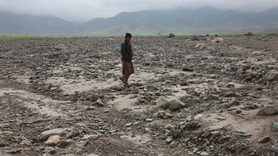A man walks through a dry, rocky riverbed in Afghanistan’s Baghlan Province, with green fields and mist-covered mountains in the background.