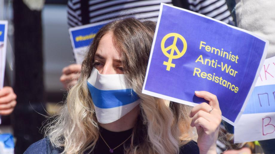 A protester wearing a face mask with a symbolic blue and white anti-war Russian flag holds a Feminist Anti-war Resistance sign.