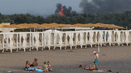 People rest on a beach as smoke and flames rise after explosions at a Russian military airbase, in Novofedorivka